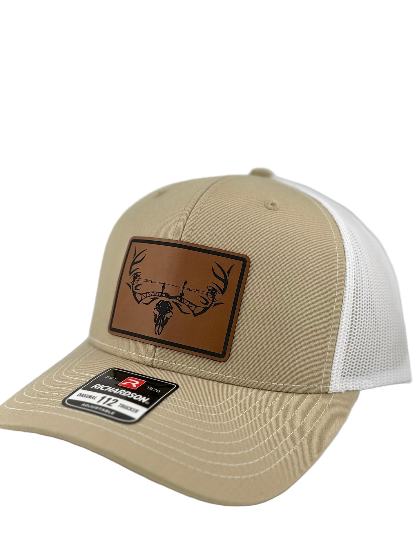 A side-angle shot of a tan and white Richardson 112 trucker hat with a brown leather patch, displaying a black deer skull and barbed wire illustration. The mesh back is partially visible.