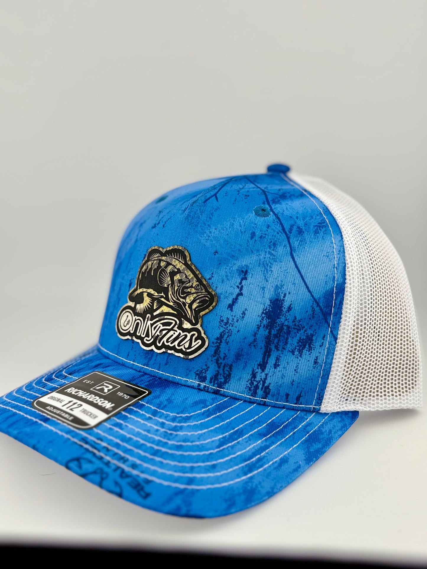 A blue and white Richardson 112 trucker hat with a water-textured pattern and a black and gold patch featuring a largemouth bass and the phrase "OnlyFins." The front panel is structured with a white mesh back.