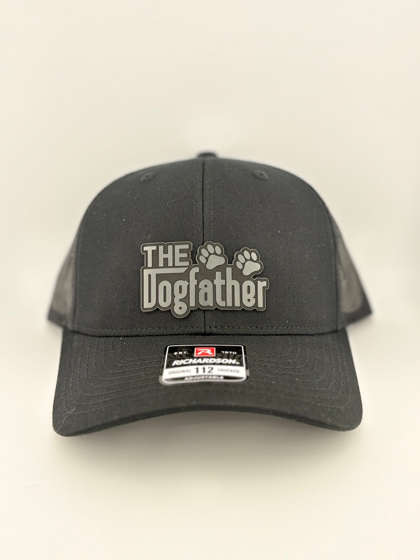 A black Richardson 112 trucker hat featuring a grey raised patch with the words "The Dogfather" and two paw prints. The hat has a structured front panel and mesh back, designed for comfort and breathability.