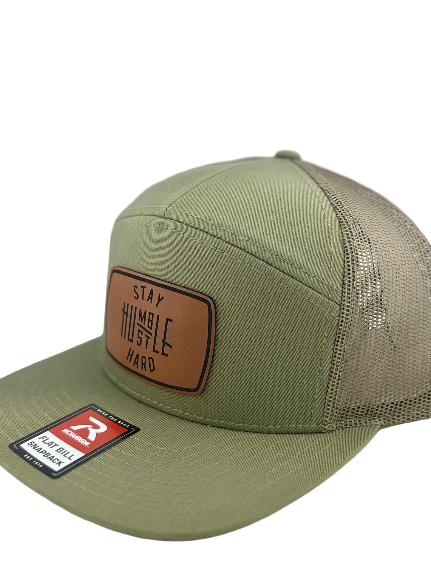 "Close-up of an olive green flat bill trucker hat with a brown leather patch displaying the phrase 'Stay Humble, Hustle Hard' in bold lettering. The hat includes a mesh back and adjustable snap closure."