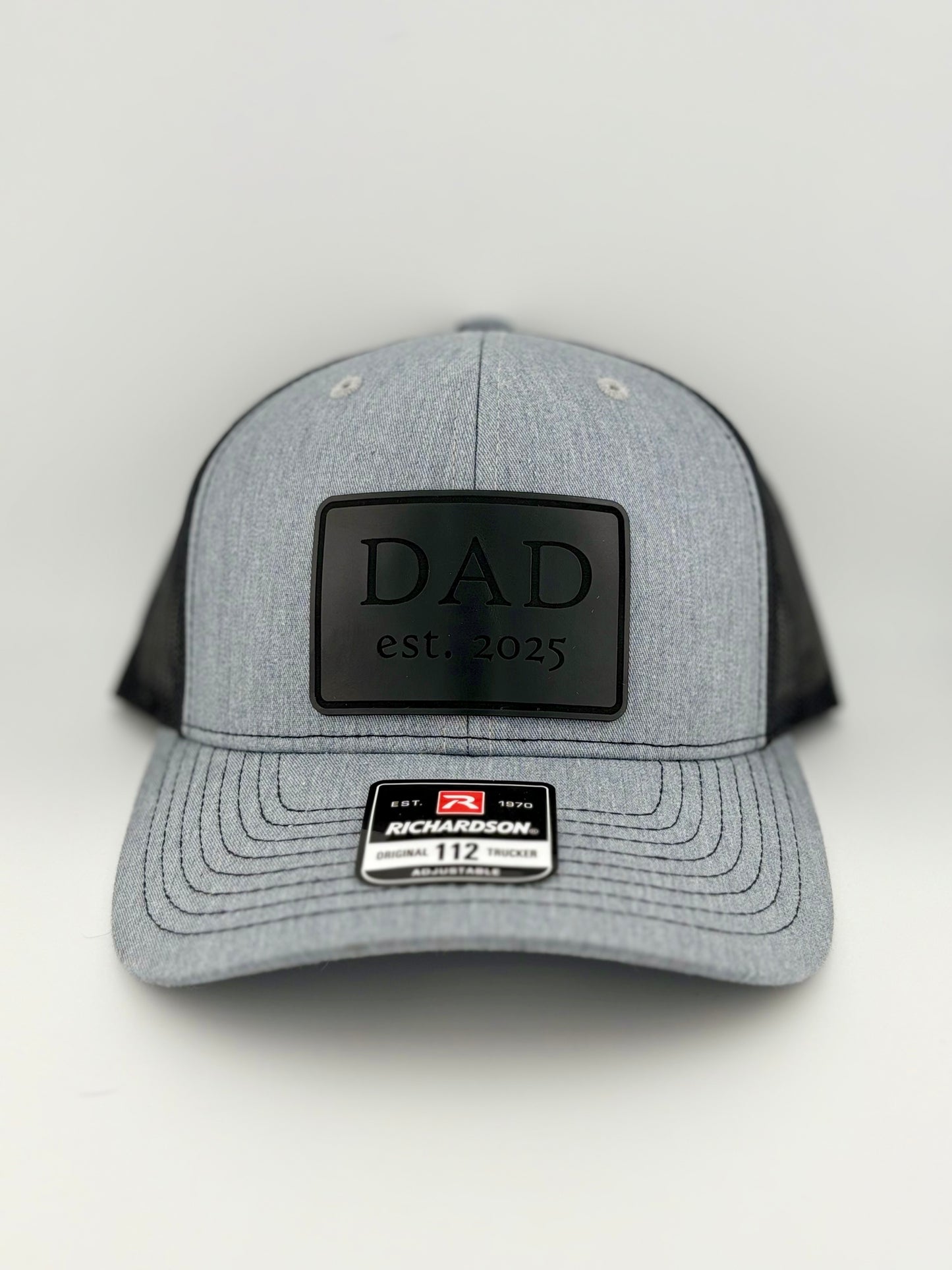 "Heather gray and black trucker hat with a structured front panel and a black mesh back. A black rectangular patch on the front features the embossed word 'DAD' in bold capital letters, followed by 'est. 2025,' making it a perfect gift for new or expecting fathers. A Richardson 112 label is affixed to the brim, indicating its adjustable fit."