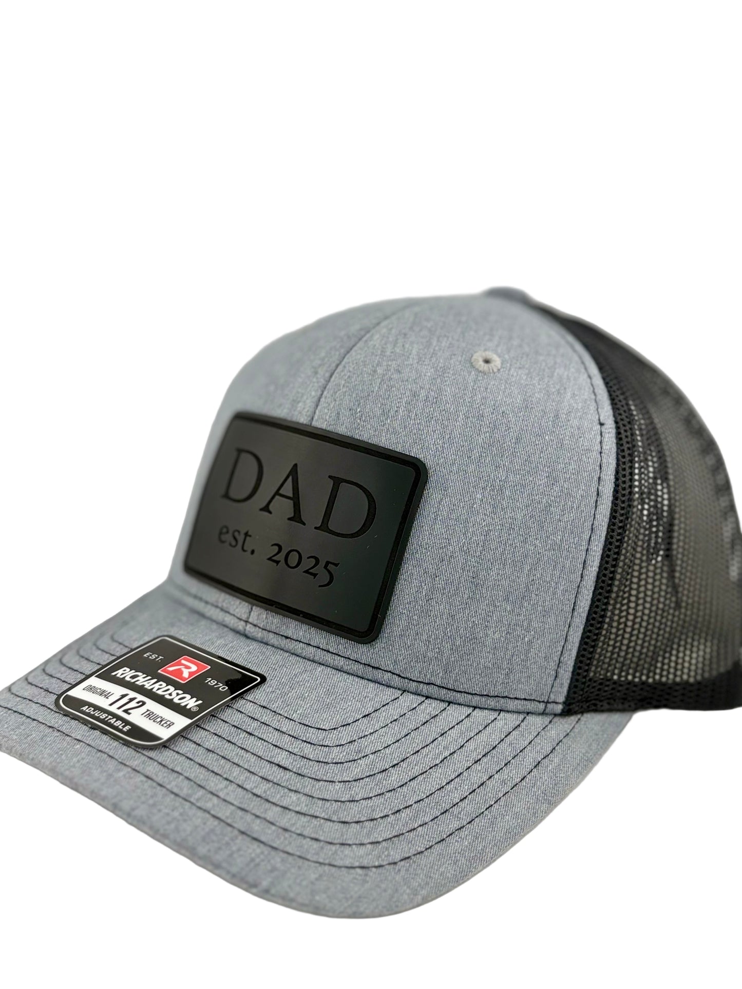 "Side view of a heather gray trucker hat with a black mesh back and structured front panel. The black rectangular patch with 'DAD est. 2025' gives the hat a sleek and sentimental look, ideal for first-time dads. A Richardson 112 label is visible on the brim."
