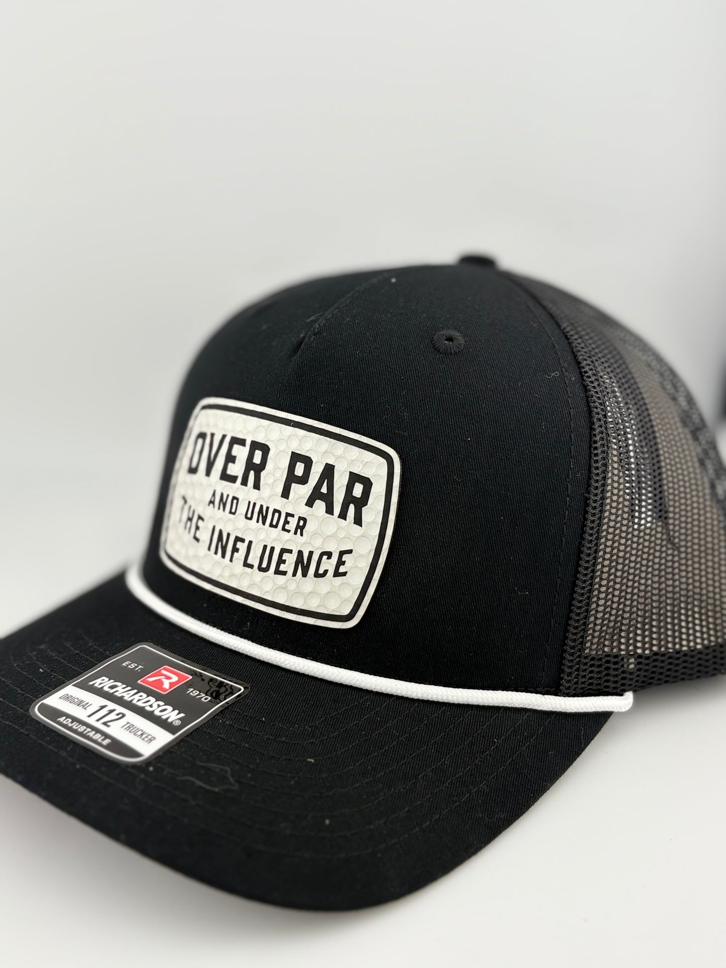 "Side view of a black trucker hat with a black mesh back and structured front panel. The white rectangular patch with a dimpled golf ball texture and the phrase 'OVER PAR AND UNDER THE INFLUENCE' adds a playful golf-themed touch. A white rope detail runs along the brim, and a Richardson 112 label is visible."