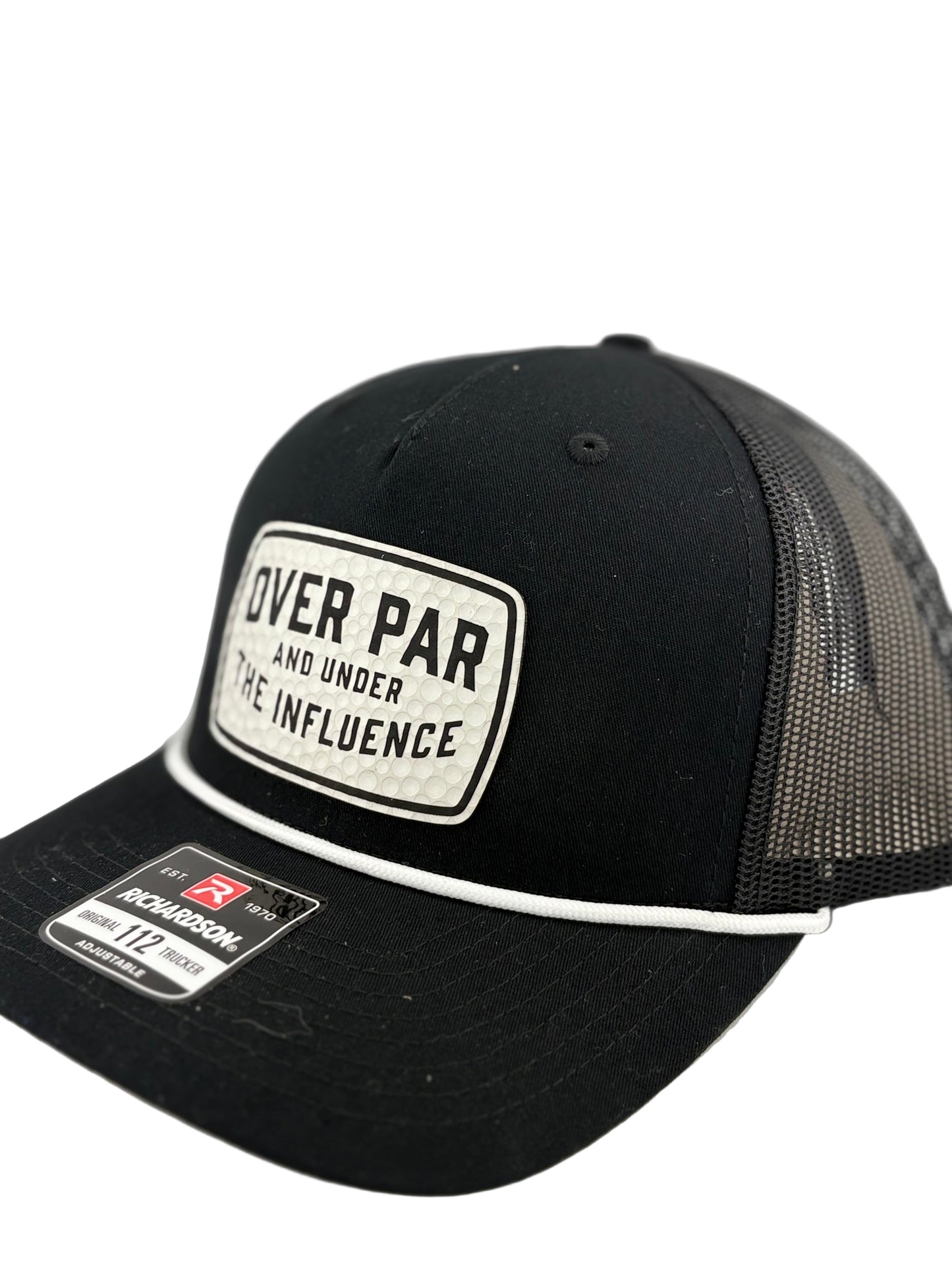 "Side view of a black trucker hat with a black mesh back and structured front panel. The white rectangular patch with a dimpled golf ball texture and the phrase 'OVER PAR AND UNDER THE INFLUENCE' adds a playful golf-themed touch. A white rope detail runs along the brim, and a Richardson 112 label is visible."