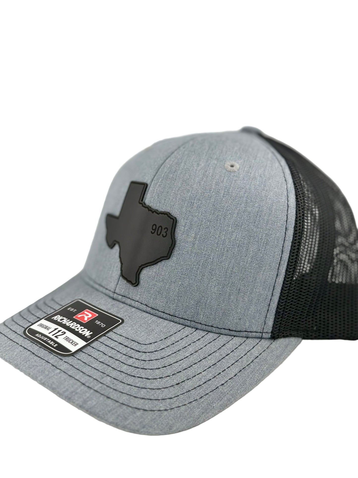 903, texas hat, 903 texas, hat, "Side view of a heather gray trucker hat with a black mesh back and structured front panel. The black Texas-shaped patch with the embossed '903' stands out, giving a bold and regional aesthetic. A Richardson 112 label is visible on the brim."