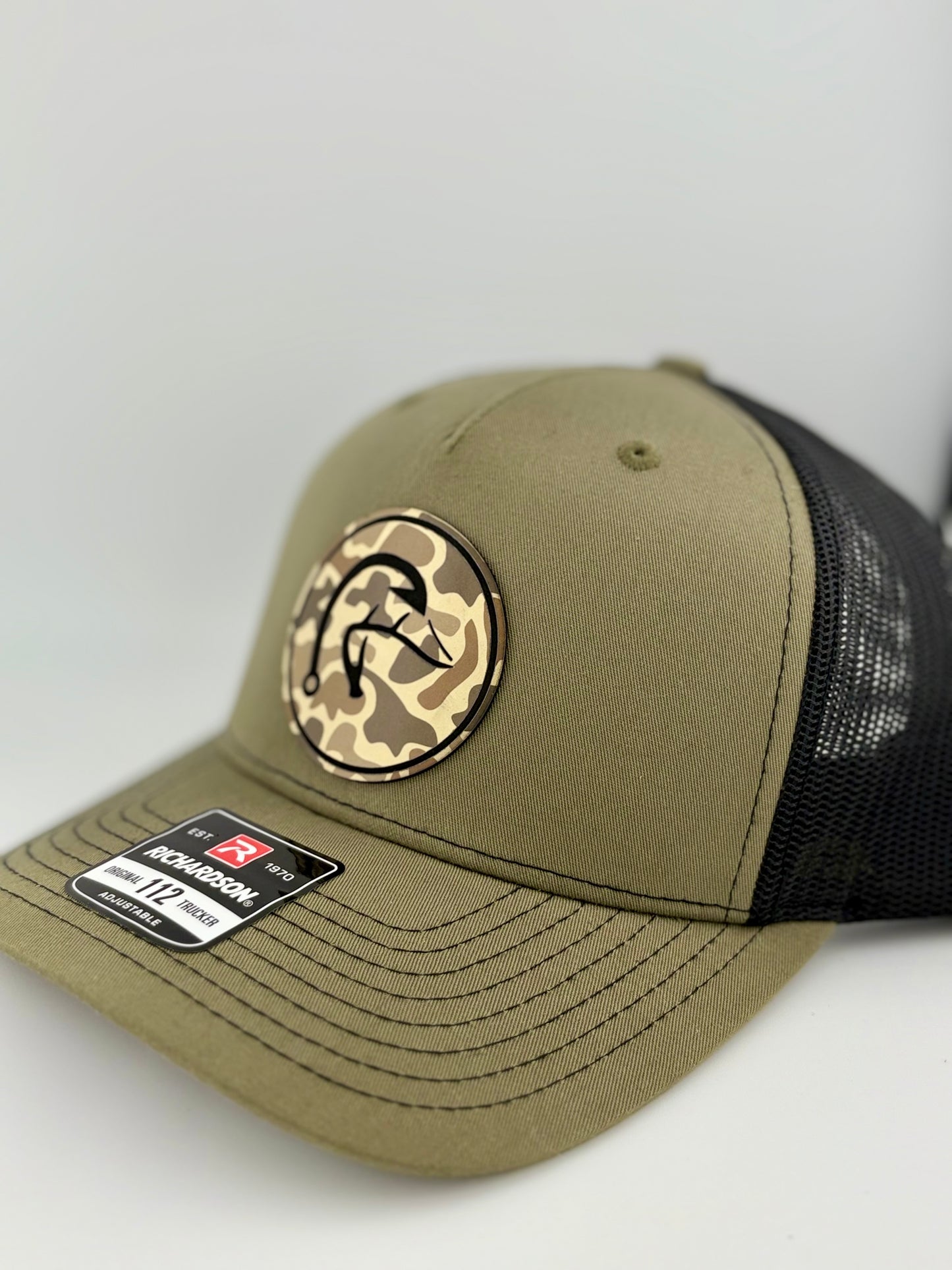 "Side view of an olive green trucker hat with a black mesh back and structured front panel. The circular patch showcases a camo background with a stylized fishhook design, adding a bold outdoor and fishing-inspired touch. A Richardson 112 label is visible on the brim."
