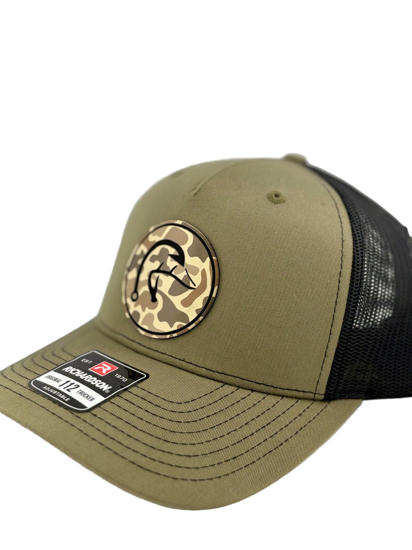 "Side view of an olive green trucker hat with a black mesh back and structured front panel. The circular patch showcases a camo background with a stylized fishhook design, adding a bold outdoor and fishing-inspired touch. A Richardson 112 label is visible on the brim."