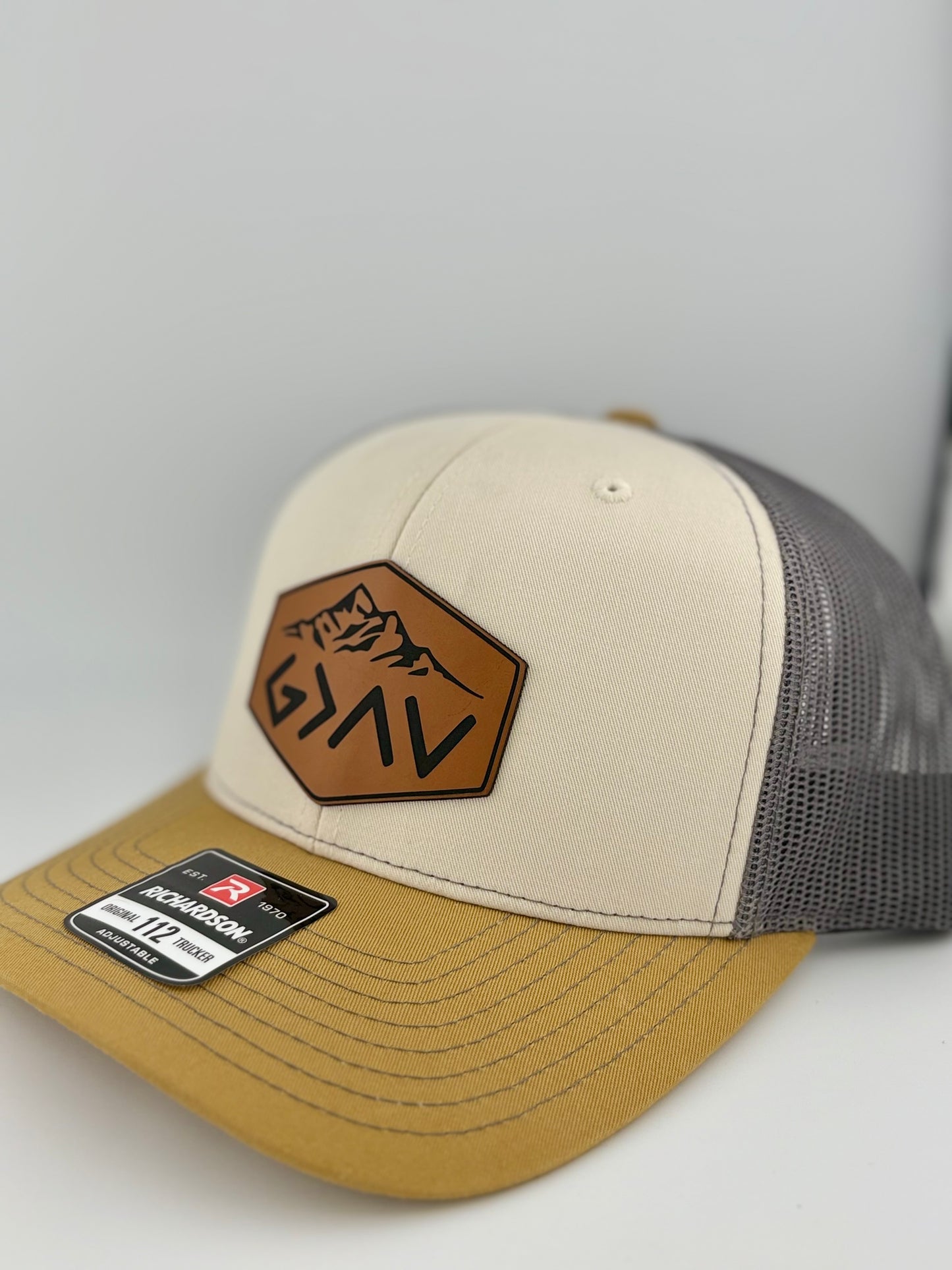 "Side view of a two-tone trucker hat with a cream front panel, mustard yellow brim, and dark gray mesh back. The brown hexagonal leather patch showcases a mountain engraving along with the 'G>^V' symbol, adding an inspirational and rugged aesthetic. A Richardson 112 label is affixed to the brim."