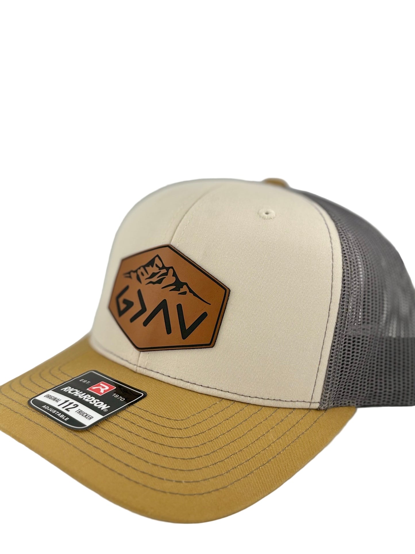 "Side view of a two-tone trucker hat with a cream front panel, mustard yellow brim, and dark gray mesh back. The brown hexagonal leather patch showcases a mountain engraving along with the 'G>^V' symbol, adding an inspirational and rugged aesthetic. A Richardson 112 label is affixed to the brim."