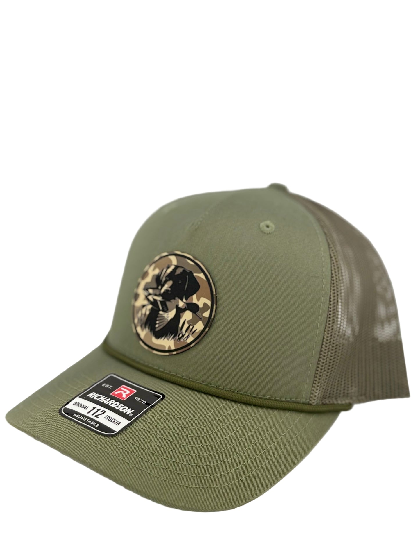 "Side view of an olive green trucker hat with a brown mesh back and structured front panel. The circular patch showcases a hunting dog with a duck in its mouth over a camo background, adding an outdoorsman’s touch. A Richardson 112 label is visible on the brim."