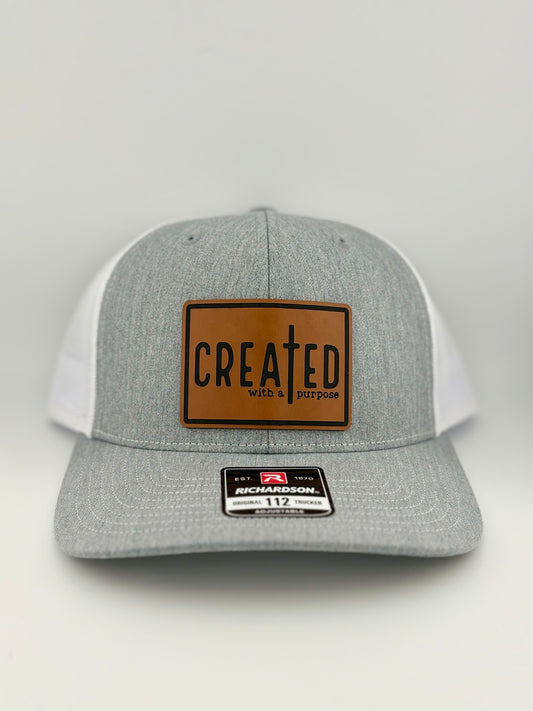 A heather gray and white trucker hat with a brown leather patch that reads "CREATED with a purpose" in bold black lettering, featuring a cross integrated into the design. The hat has a mesh back and a Richardson 112 sticker on the brim.