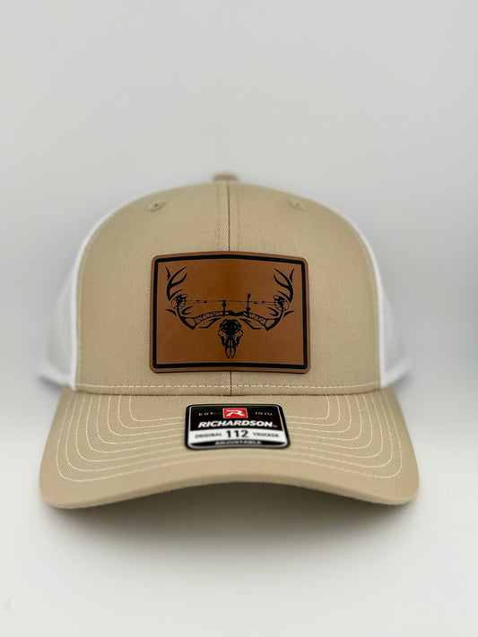 A tan and white Richardson 112 trucker hat featuring a brown leather patch with a black illustration of a deer skull with antlers integrated into a barbed wire design.