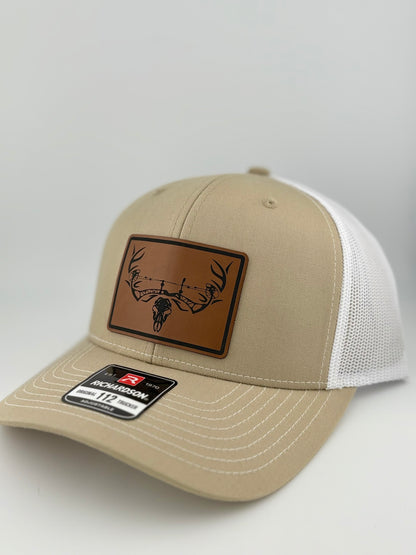 A side-angle shot of a tan and white Richardson 112 trucker hat with a brown leather patch, displaying a black deer skull and barbed wire illustration. The mesh back is partially visible.