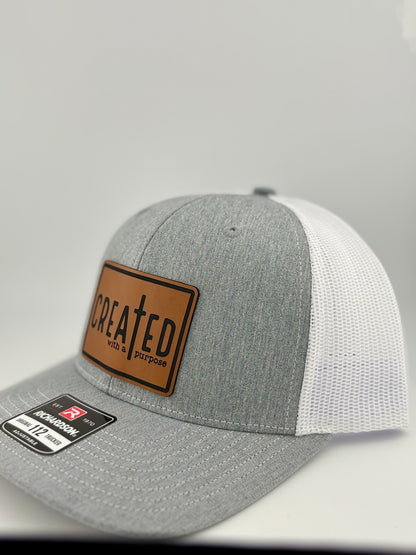 A heather gray and white trucker hat displayed at an angle, showing the mesh back and side details. The brown leather patch on the front reads "CREATED with a purpose" with a cross as part of the design. The structured front panel and adjustable fit are visible.