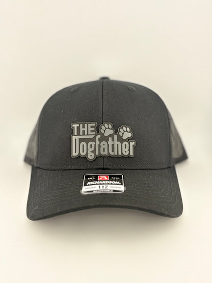 A black Richardson 112 trucker hat featuring a grey raised patch with the words "The Dogfather" and two paw prints. The hat has a structured front panel and mesh back, designed for comfort and breathability.