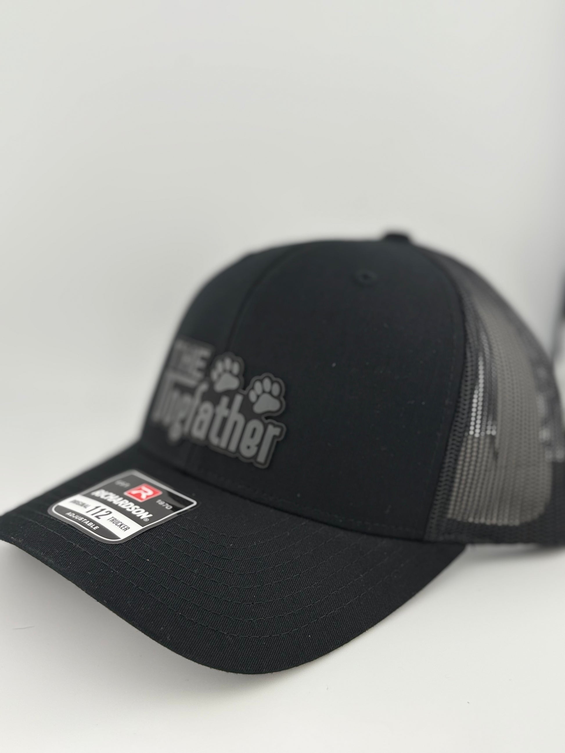 A side view of the black Richardson 112 trucker hat with "The Dogfather" patch, showcasing the 3D effect of the lettering and paw prints. The hat has a curved bill and mesh back for a classic trucker style.