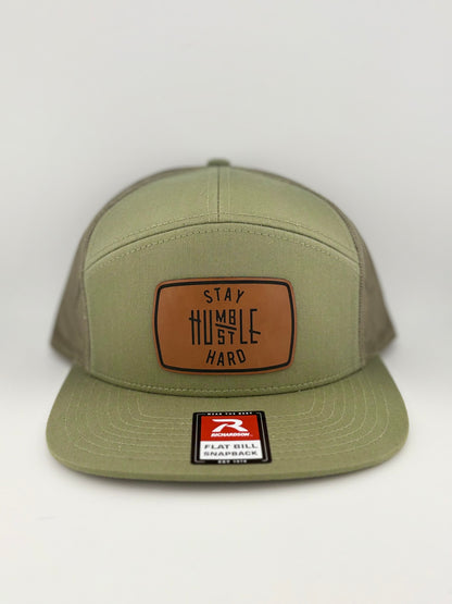 "Olive green Richardson 112 snapback hat with a leather patch that reads 'Stay Humble, Hustle Hard.' The hat features a structured front panel, mesh back, and a flat bill."