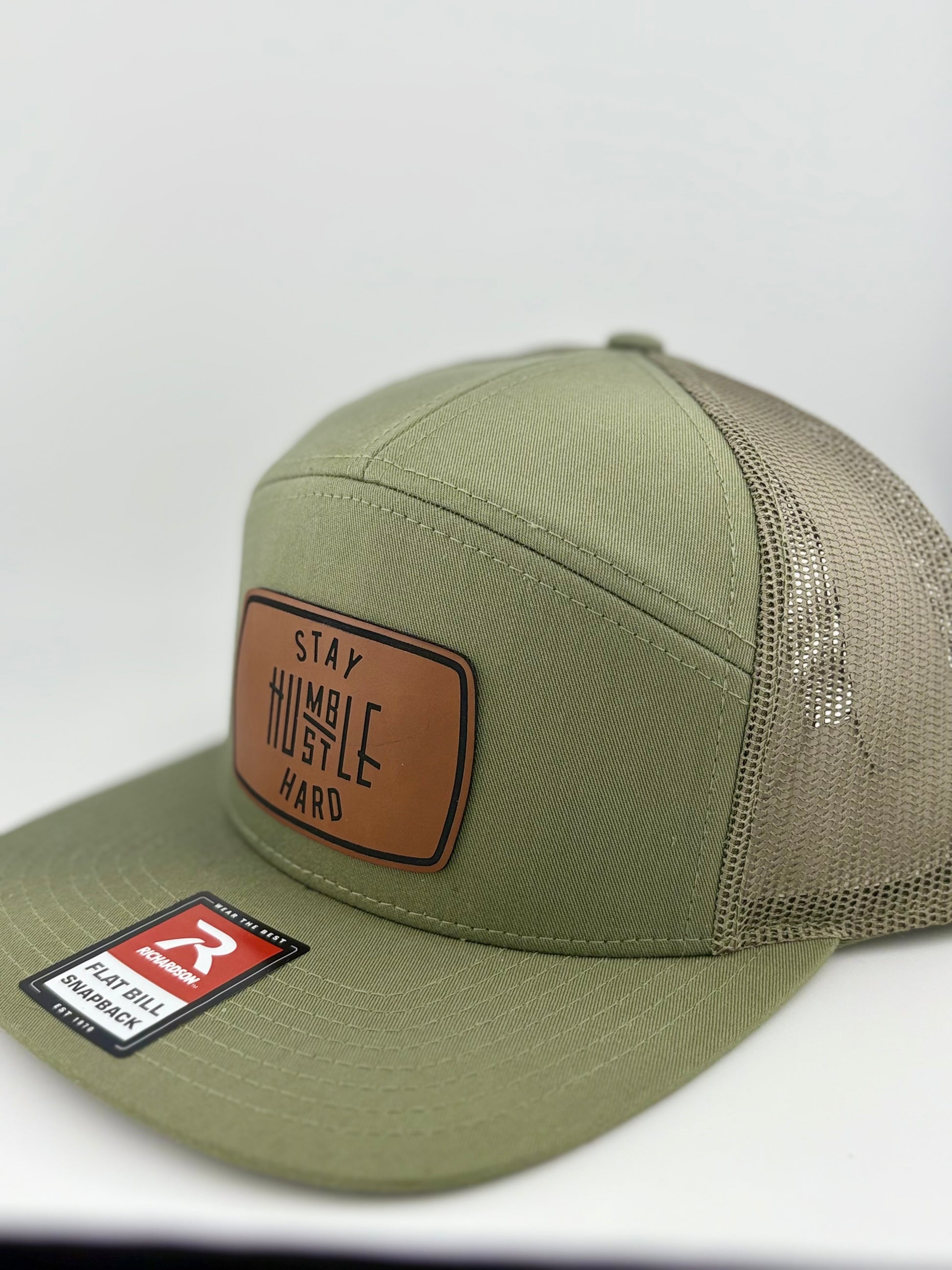 "Close-up of an olive green flat bill trucker hat with a brown leather patch displaying the phrase 'Stay Humble, Hustle Hard' in bold lettering. The hat includes a mesh back and adjustable snap closure."