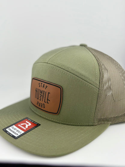 "Close-up of an olive green flat bill trucker hat with a brown leather patch displaying the phrase 'Stay Humble, Hustle Hard' in bold lettering. The hat includes a mesh back and adjustable snap closure."