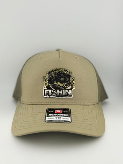 "Tan trucker hat with a structured front panel and a tan mesh back. The front features a detailed patch with a stylized bass fish wearing sunglasses, surrounded by the phrase 'WISHIN' I WAS FISHIN'.' The bold, metallic design makes it a standout for fishing enthusiasts. A Richardson 112 label is affixed to the brim, indicating an adjustable fit."