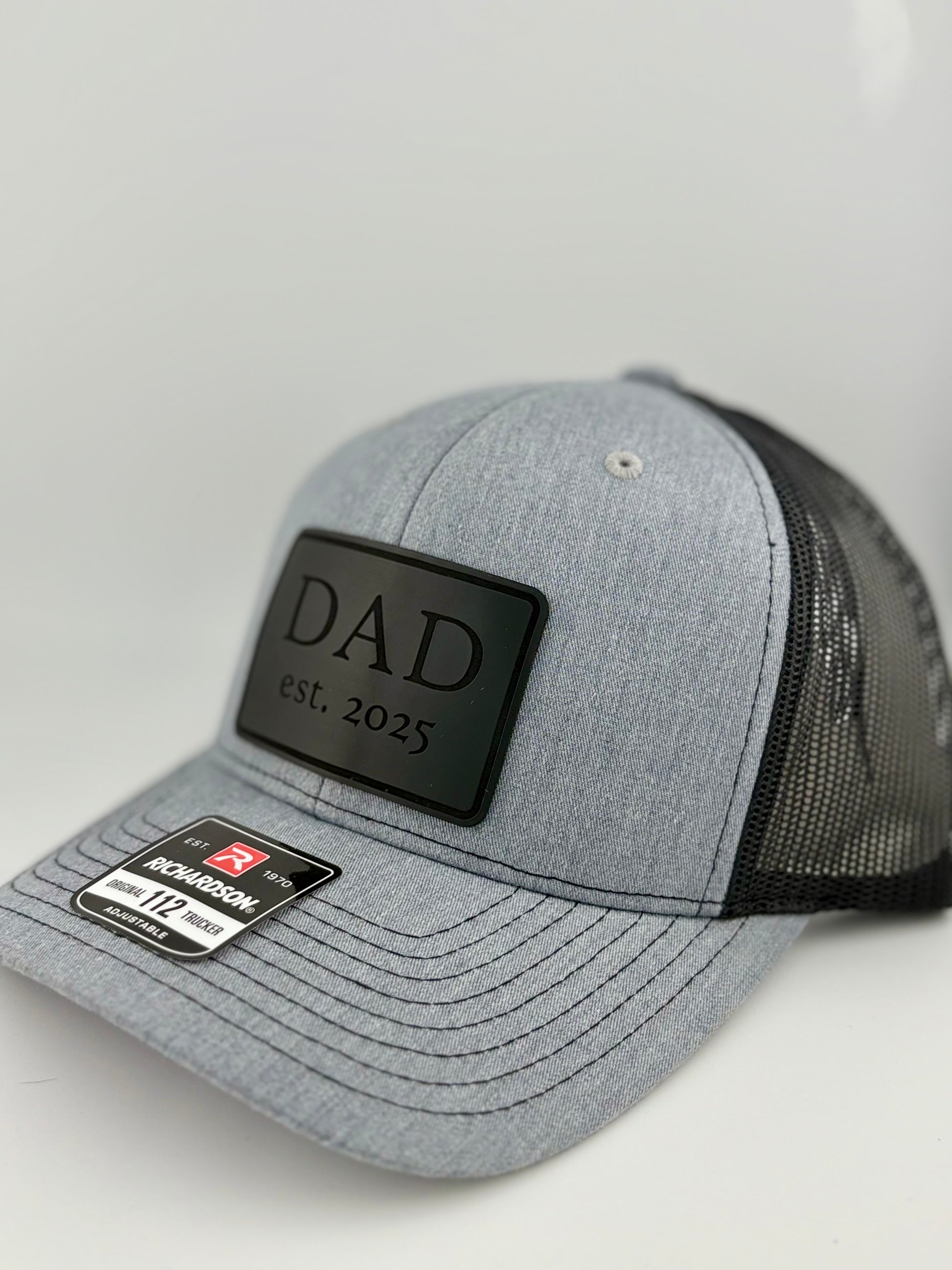 "Side view of a heather gray trucker hat with a black mesh back and structured front panel. The black rectangular patch with 'DAD est. 2025' gives the hat a sleek and sentimental look, ideal for first-time dads. A Richardson 112 label is visible on the brim."