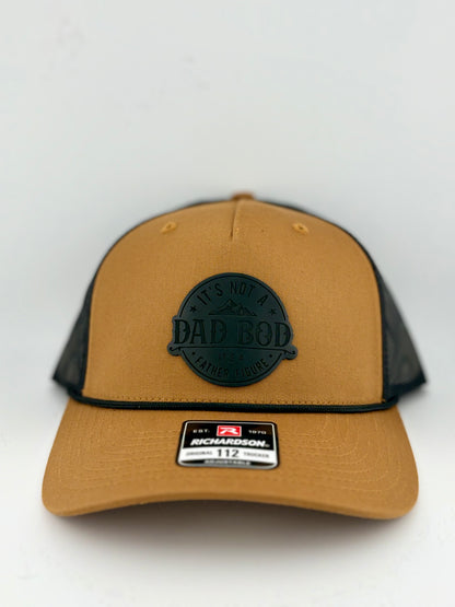 "Tan and black trucker hat with a structured front panel and a black mesh back. A black circular patch on the front features the phrase 'IT'S NOT A DAD BOD, IT'S A FATHER FIGURE' along with a mountain design. A black rope detail runs along the brim, and a Richardson 112 label is affixed to the front."