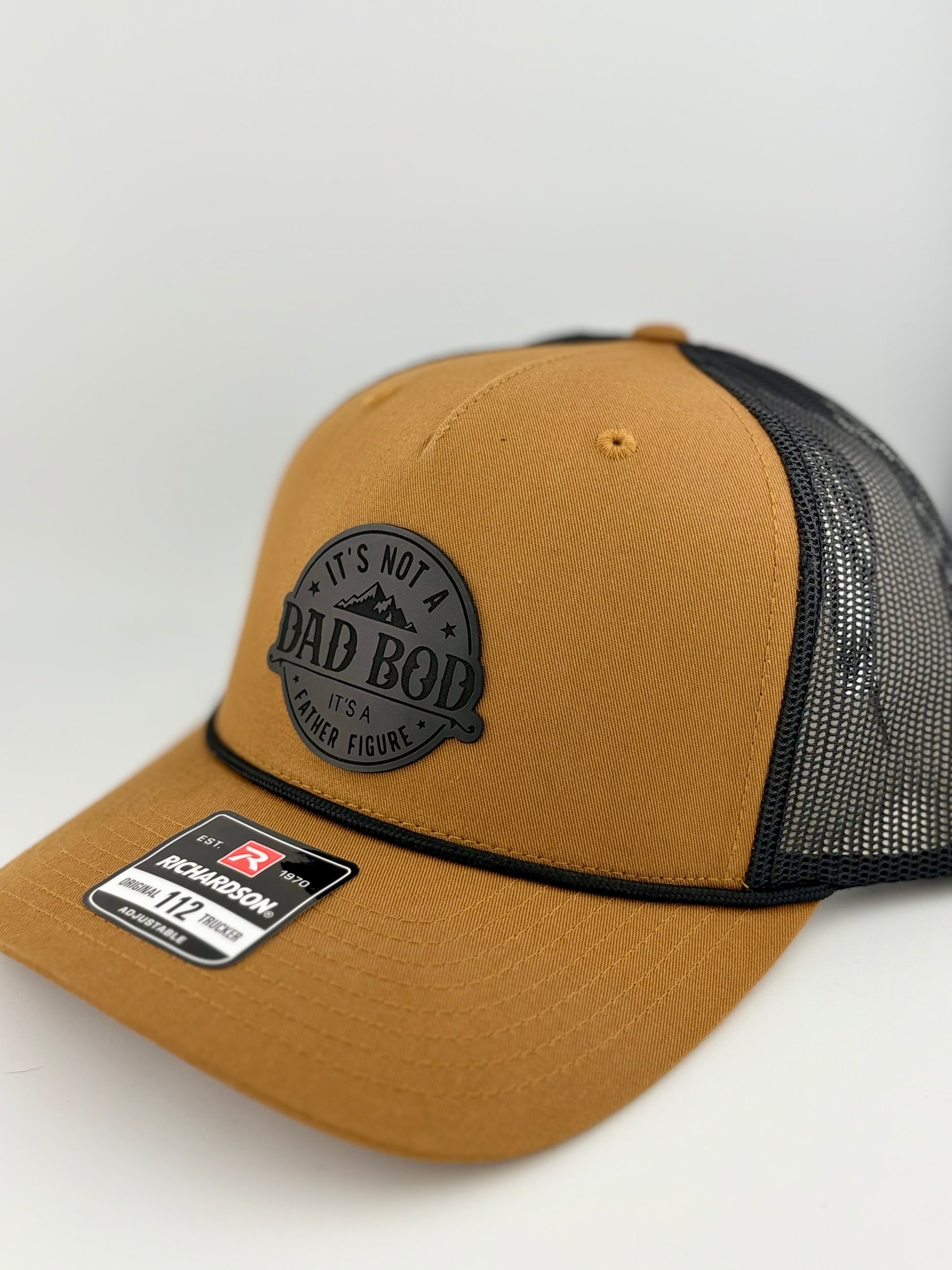 "Side view of a tan trucker hat with a black mesh back and structured front panel. The black circular patch with 'IT'S NOT A DAD BOD, IT'S A FATHER FIGURE' and a mountain design gives the hat a rugged and humorous touch. A black rope detail is featured along the brim, and a Richardson 112 label is visible."