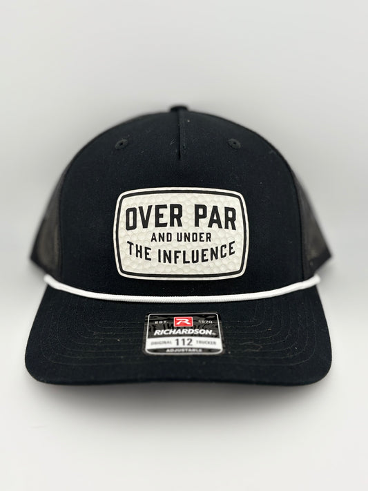 "Black trucker hat with a structured front panel and a black mesh back. A white rectangular patch on the front features a dimpled golf ball texture with the humorous phrase 'OVER PAR AND UNDER THE INFLUENCE' in bold black capital letters. A white rope detail runs across the brim, and a Richardson 112 label is affixed to the front."