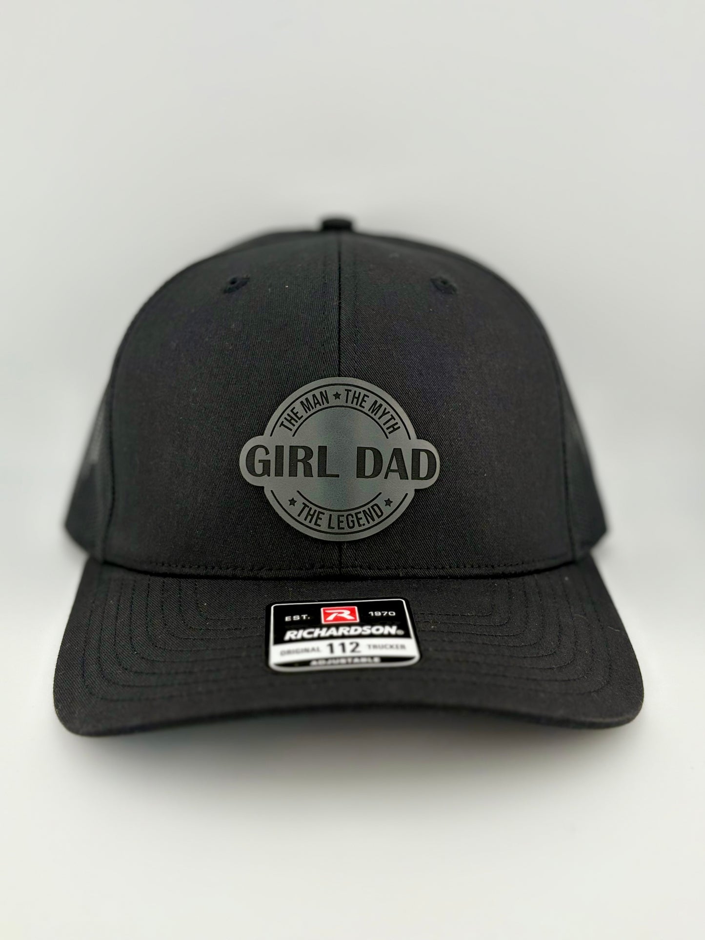 "Black trucker hat with a structured front panel and black mesh back. A circular gray patch on the front features the words 'GIRL DAD' in bold capital letters, surrounded by the phrases 'The Man • The Myth • The Legend.' A Richardson 112 label is affixed to the brim, indicating its adjustable fit."