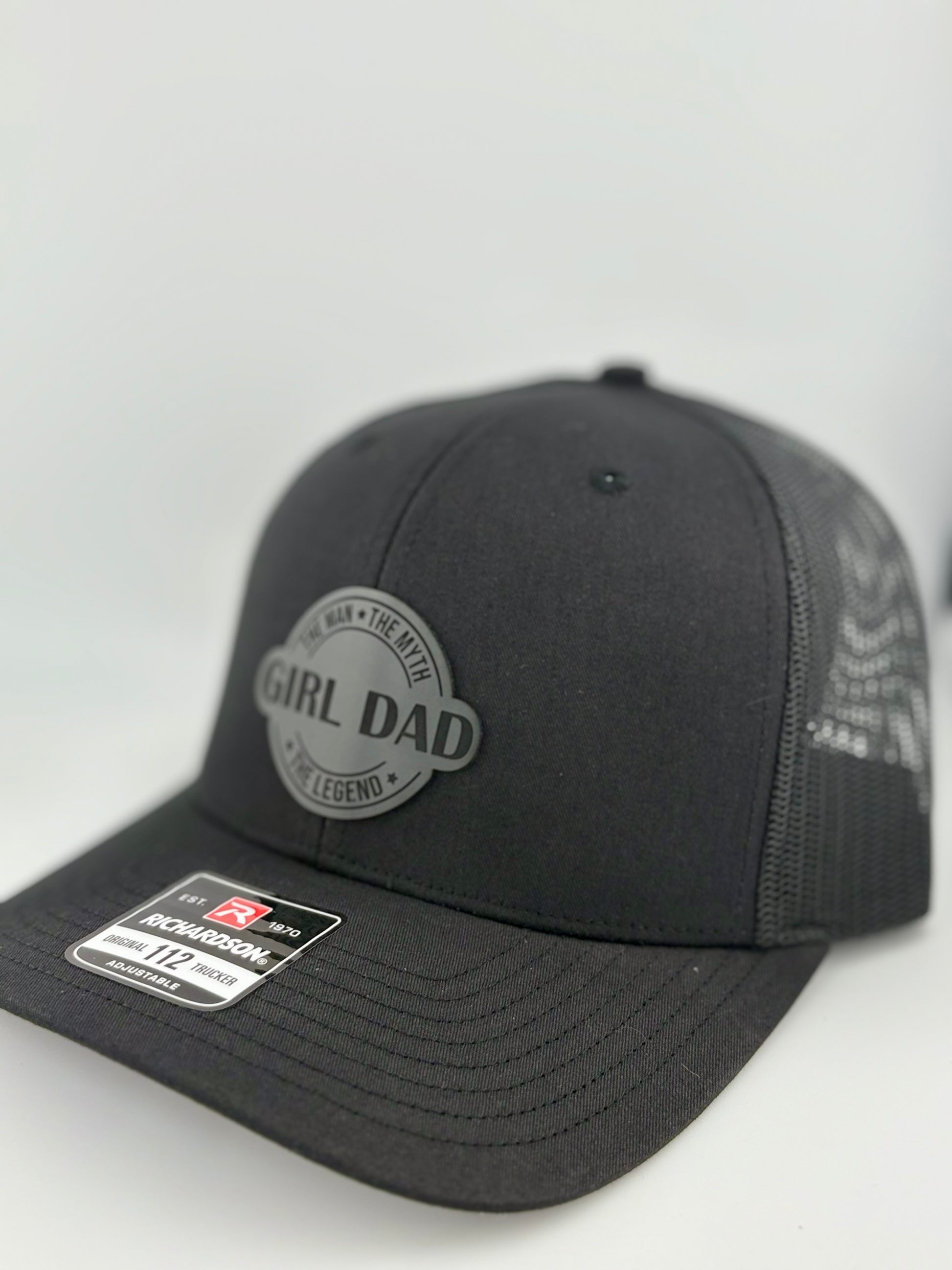 "Side view of a black trucker hat with a black mesh back and structured front panel. The circular gray patch with 'GIRL DAD' and the phrases 'The Man • The Myth • The Legend' gives the hat a sleek, modern, and proud fatherhood aesthetic. A Richardson 112 label is visible on the brim."