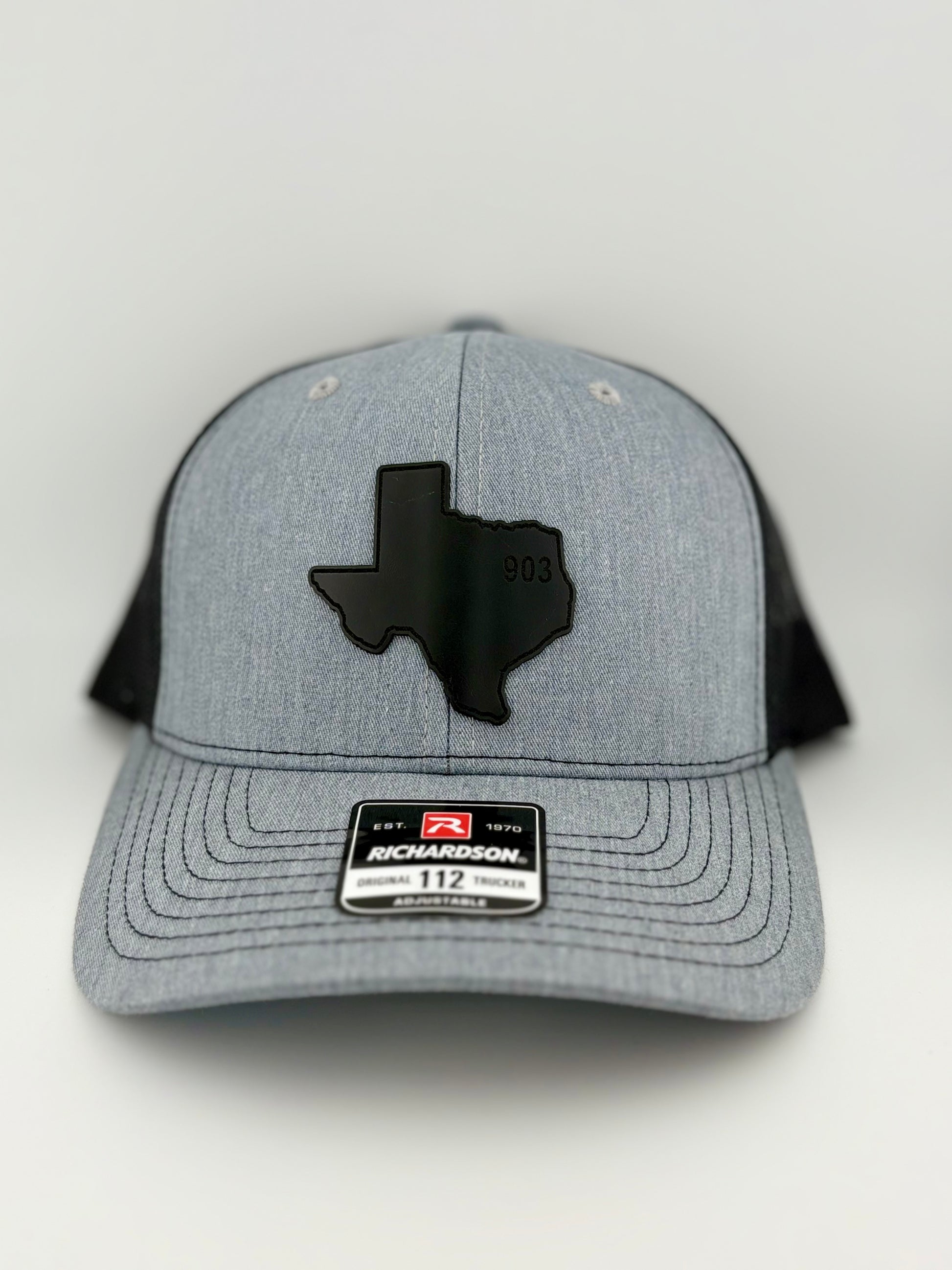 903, texas hat, 903 texas, hat, "Heather gray and black trucker hat with a structured front panel and a black mesh back. A black Texas-shaped patch is featured on the front, embossed with the number '903,' representing the East Texas area code. A Richardson 112 label is affixed to the brim, indicating its adjustable fit."