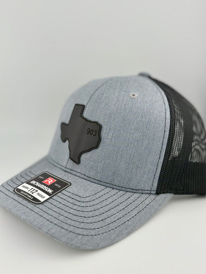 903, texas hat, 903 texas, hat, "Side view of a heather gray trucker hat with a black mesh back and structured front panel. The black Texas-shaped patch with the embossed '903' stands out, giving a bold and regional aesthetic. A Richardson 112 label is visible on the brim."