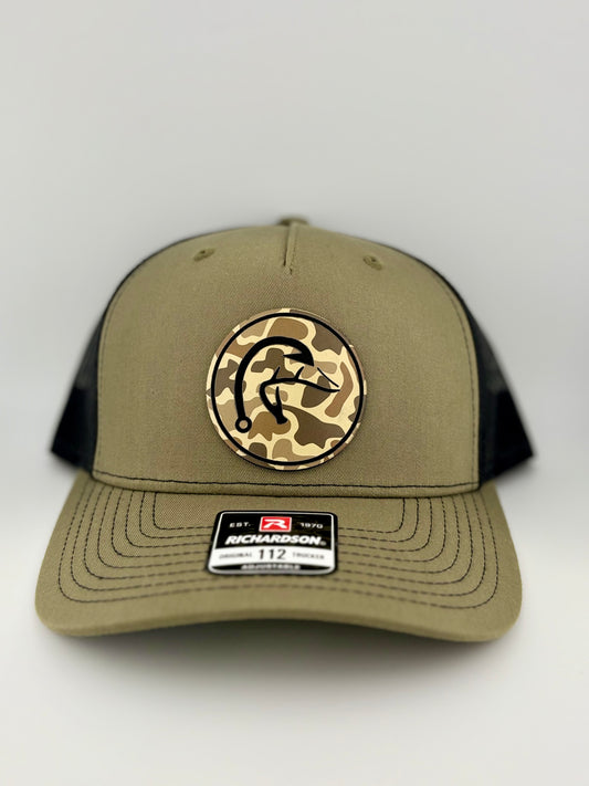 "Olive green and black trucker hat with a structured front panel and a mesh back. A circular patch on the front features a camo background with an engraved stylized fishhook design. A Richardson 112 label is affixed to the brim, indicating its adjustable fit."