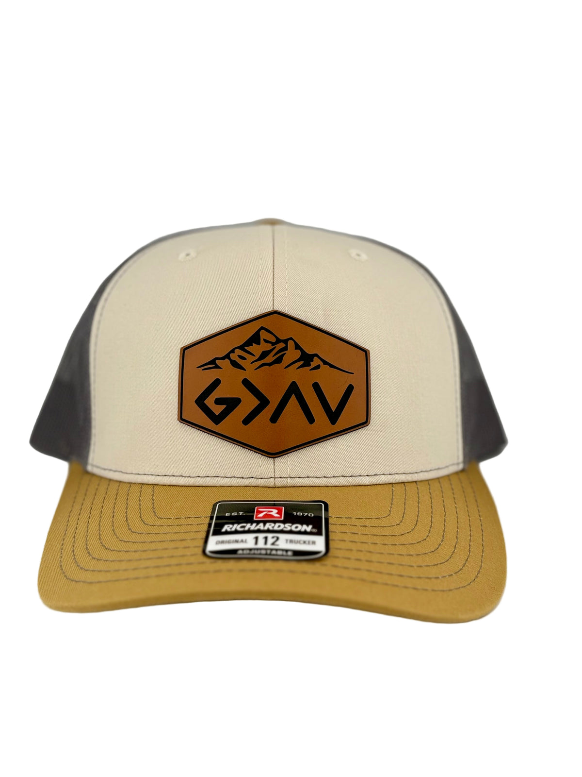 "Two-tone trucker hat with a cream-colored front panel, mustard yellow brim, and dark gray mesh back. A brown hexagonal leather patch on the front features an engraved mountain design and the faith-based symbol 'G>^V' (God is greater than the highs and lows). A Richardson 112 label is attached to the brim."