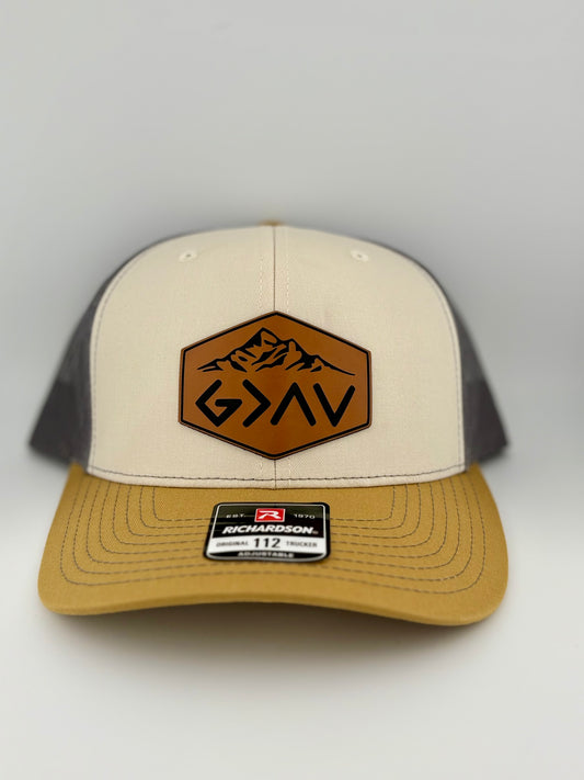 "Two-tone trucker hat with a cream-colored front panel, mustard yellow brim, and dark gray mesh back. A brown hexagonal leather patch on the front features an engraved mountain design and the faith-based symbol 'G>^V' (God is greater than the highs and lows). A Richardson 112 label is attached to the brim."