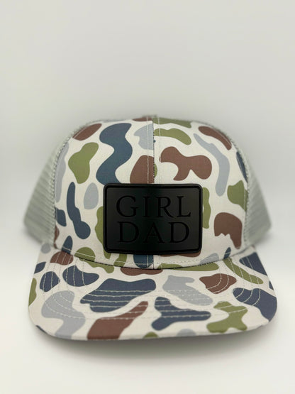 "Camo-patterned trucker hat with a structured front panel and a light gray mesh back. A black rectangular patch on the front features the embossed words 'GIRL DAD' in bold capital letters, creating a stylish and modern tribute to fatherhood."