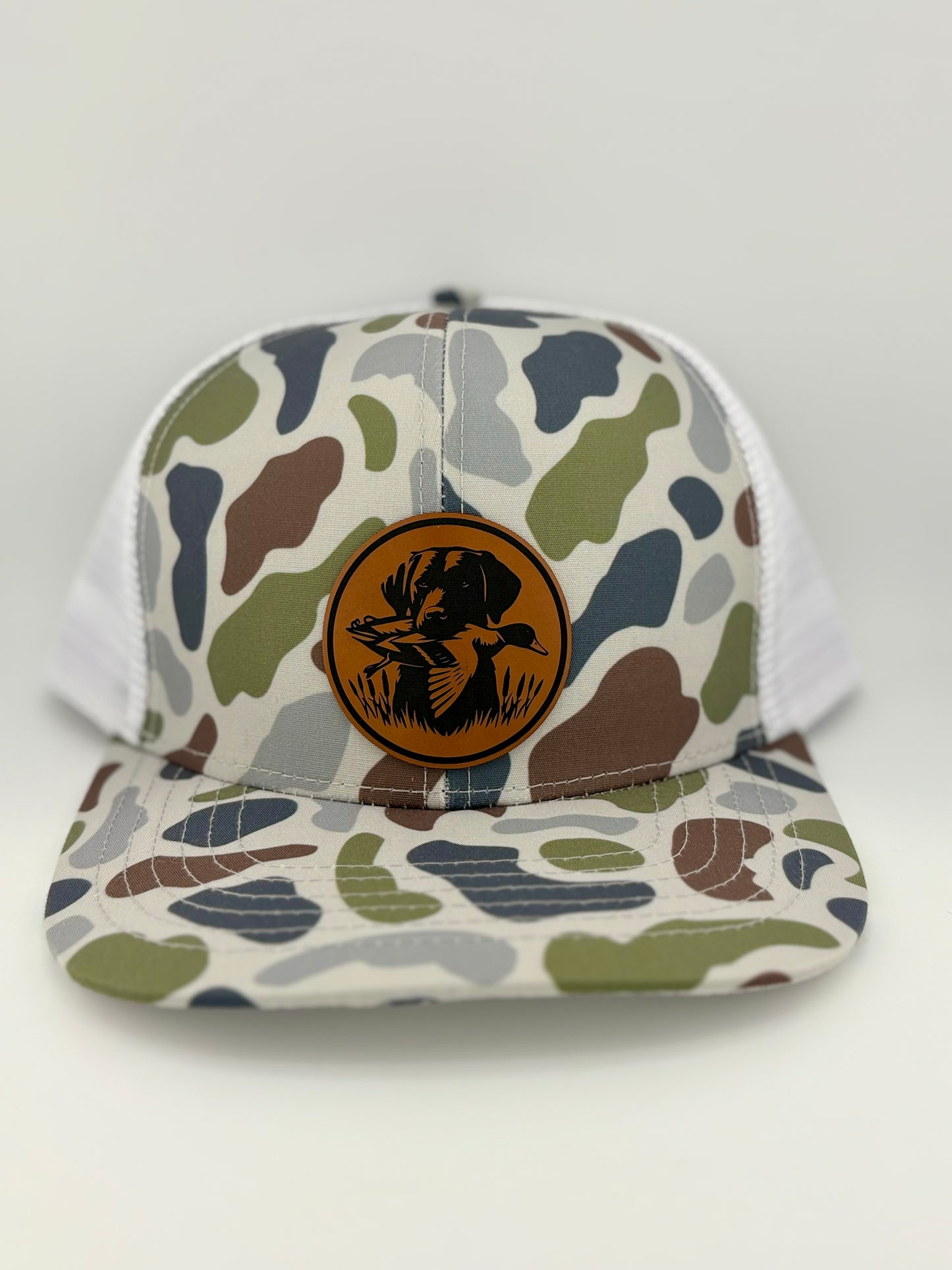 "Camo-patterned trucker hat with a structured front panel and a white mesh back. A brown leather patch on the front features an engraved hunting dog holding a duck in its mouth, set within a circular border, embodying a classic waterfowl hunting theme."