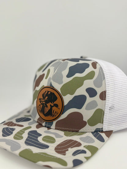 "Side view of a camo trucker hat with a white mesh back and a structured front panel. The brown leather patch displays a hunting dog retrieving a duck, creating a rugged and outdoorsy aesthetic perfect for hunting enthusiasts."