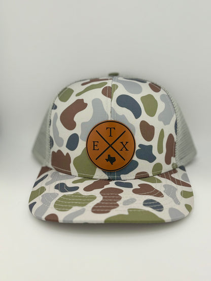 "Camo-patterned trucker hat with a structured front panel and a light gray mesh back. A brown leather patch on the front displays a circular design with crossed lines, the letters 'E,' 'T,' and 'X,' and an engraved Texas state outline, representing East Texas pride."