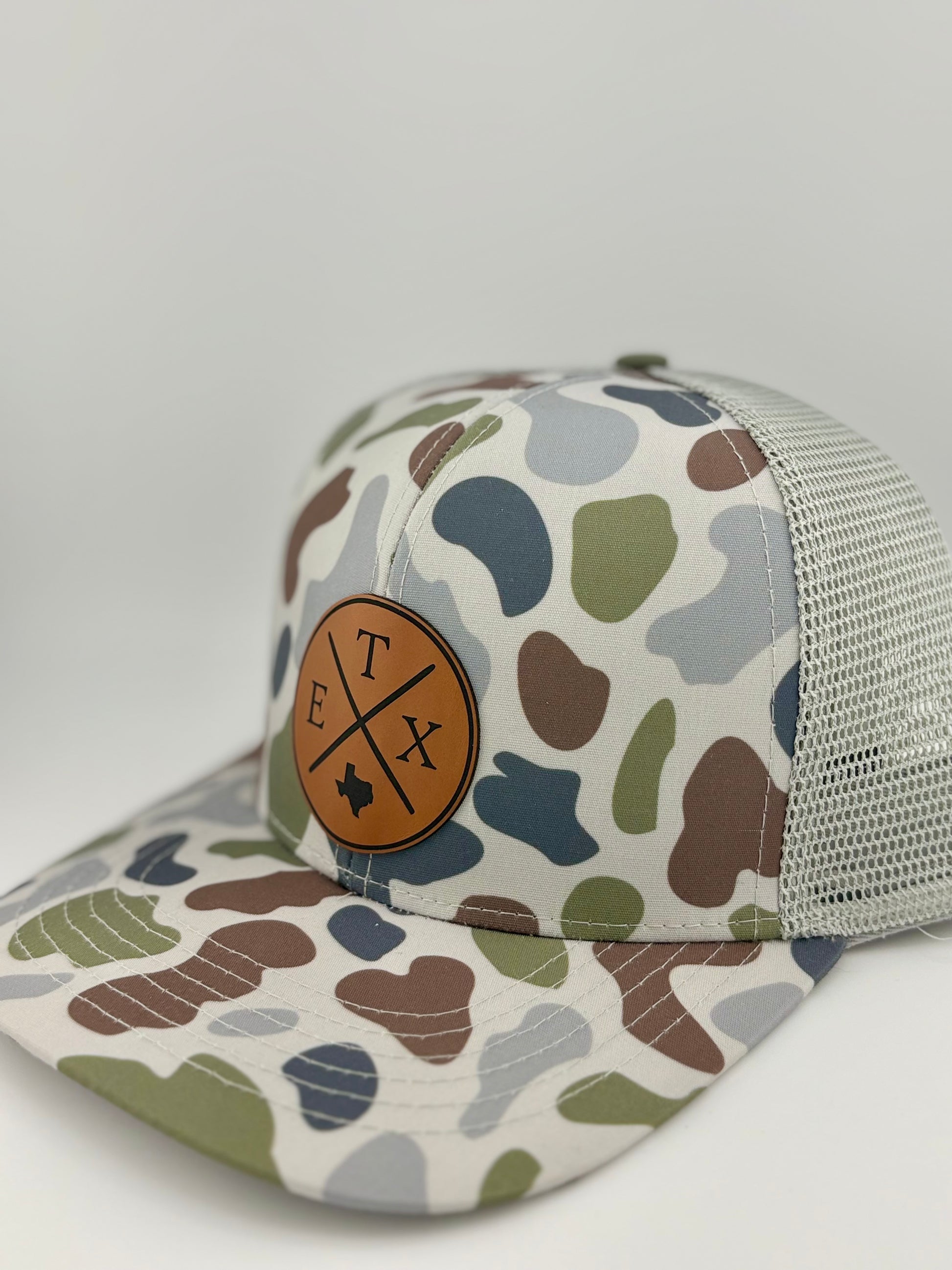 "Side view of a camo trucker hat with a light gray mesh back and a structured front panel. The brown leather patch features a crossed-line design with 'E,' 'T,' and 'X,' along with a Texas state silhouette, making it a stylish and regional statement piece."