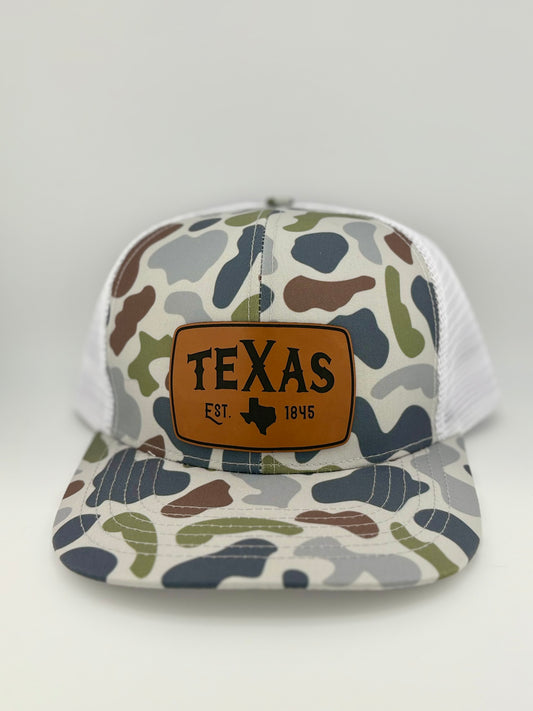 "Camo-patterned trucker hat with a structured front panel and a white mesh back. A brown leather patch on the front features the word 'TEXAS' in bold, capital letters, along with the outline of the state and 'EST. 1845,' commemorating Texas' founding year."