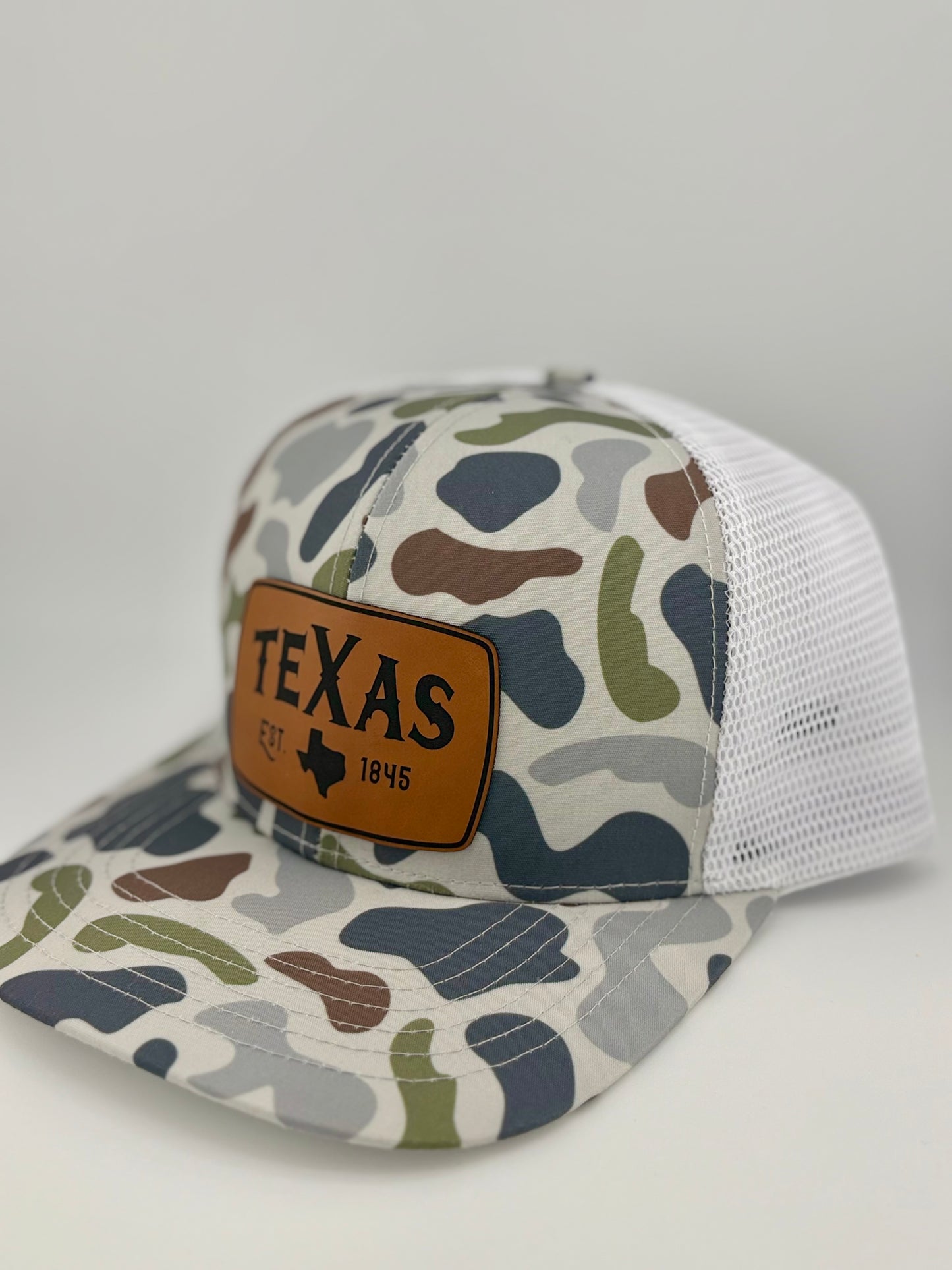 "Side view of a camo trucker hat with a white mesh back and a structured front panel. The brown leather patch displays 'TEXAS,' the state outline, and 'EST. 1845,' creating a rustic and outdoorsy look that celebrates Texas pride."