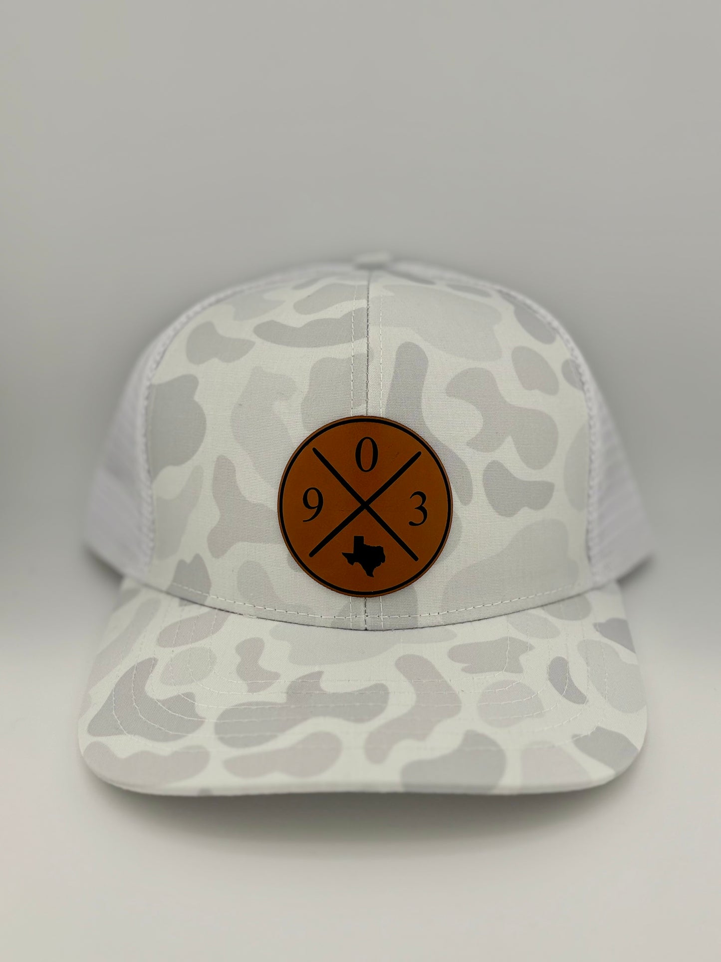 "White and light gray camo trucker hat with a structured front panel and a white mesh back. A brown leather patch on the front features a circular design with crossed lines, the numbers '9', '0' and '3,' and an engraved outline of Texas, representing the 903 area code."