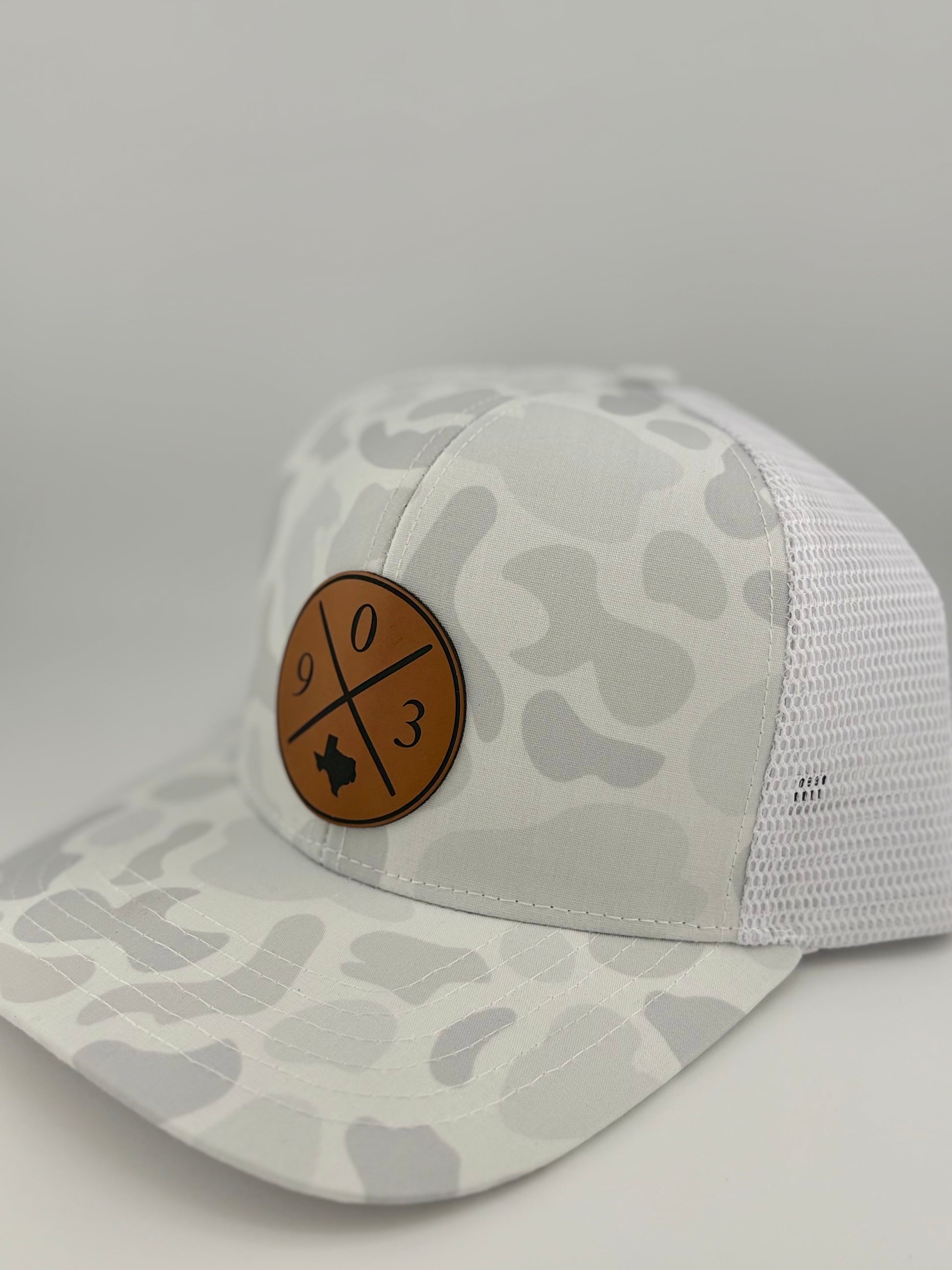 "Side view of a white and light gray camo-patterned trucker hat with a structured front and white mesh back. The brown leather patch on the front displays a crossed-line design with '9', '0' and '3,' along with a Texas state silhouette, adding a regional touch to the stylish headwear."