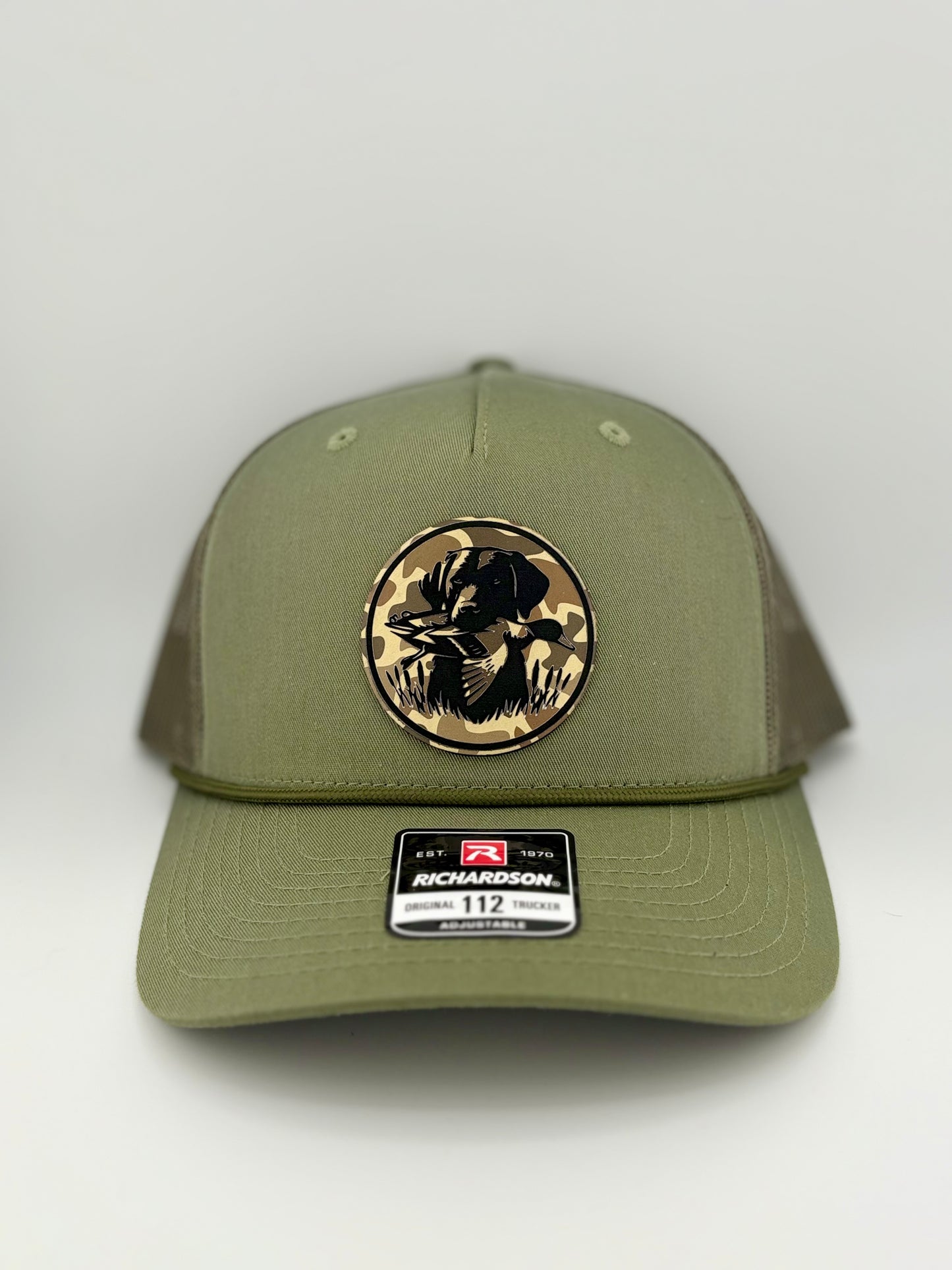 "Olive green and brown trucker hat with a structured front panel and a mesh back. The front features a circular patch with a camo background and an engraved hunting dog holding a duck, symbolizing a strong waterfowl hunting tradition. A Richardson 112 label is placed on the brim."