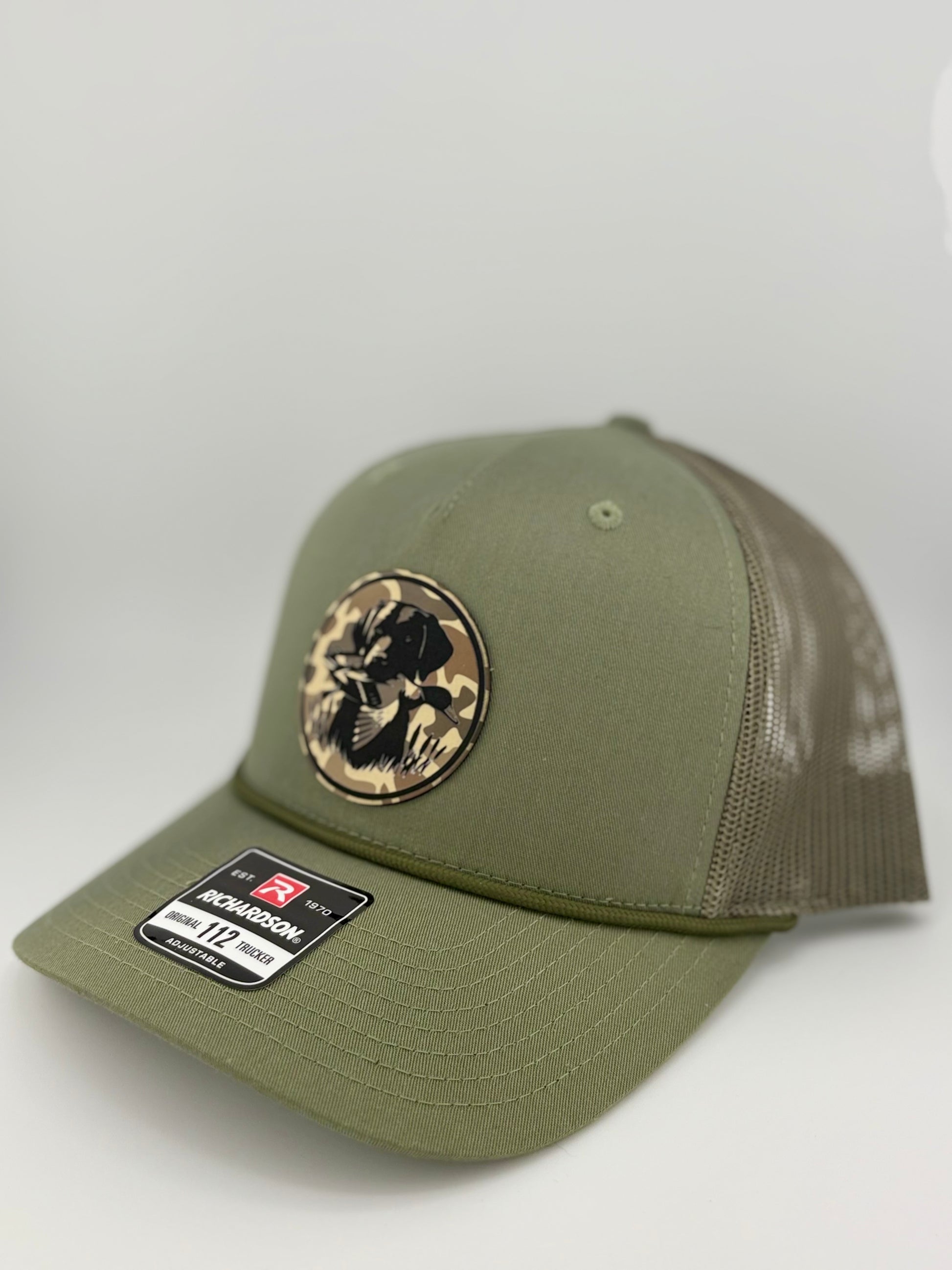 "Side view of an olive green trucker hat with a brown mesh back and structured front panel. The circular patch showcases a hunting dog with a duck in its mouth over a camo background, adding an outdoorsman’s touch. A Richardson 112 label is visible on the brim."