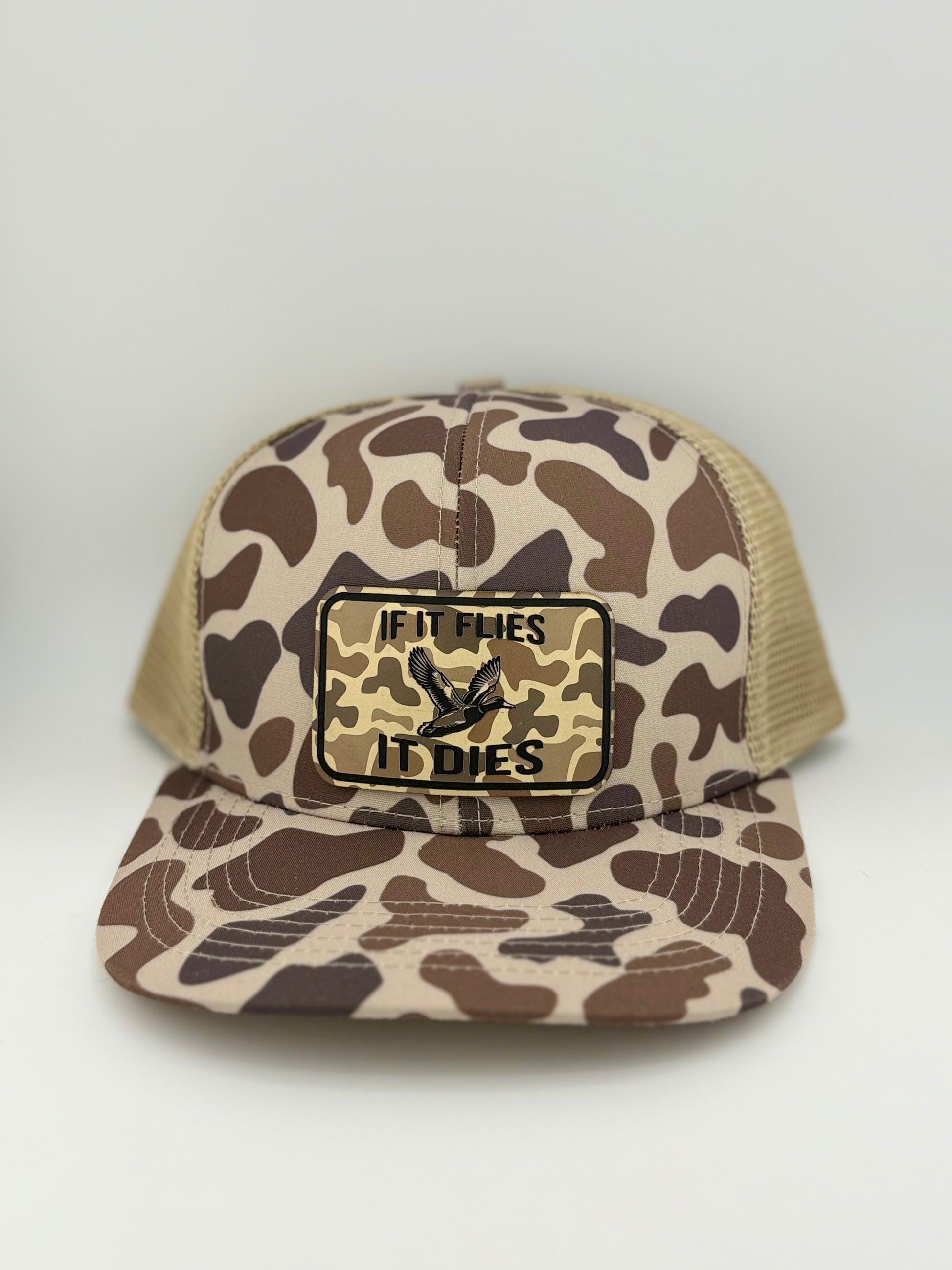 "Brown and tan camo trucker hat with a structured front panel and a breathable mesh back. The front features a rectangular patch with a matching camo background, an illustration of a flying duck, and the bold phrase 'IF IT FLIES IT DIES' in black capital letters, catering to outdoor and hunting enthusiasts."