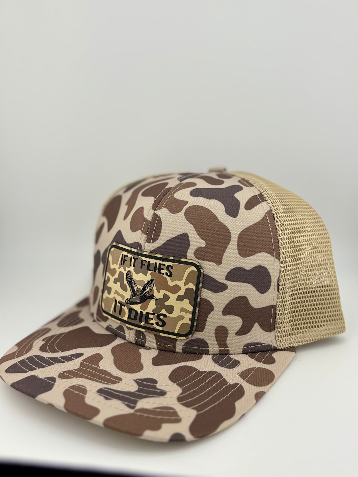 "Side view of a brown and tan camo-patterned trucker hat with a structured front and tan mesh back. The front patch, featuring a flying duck and the phrase 'IF IT FLIES IT DIES,' stands out against the camo backdrop, making it a bold statement piece for hunters and outdoor lovers."