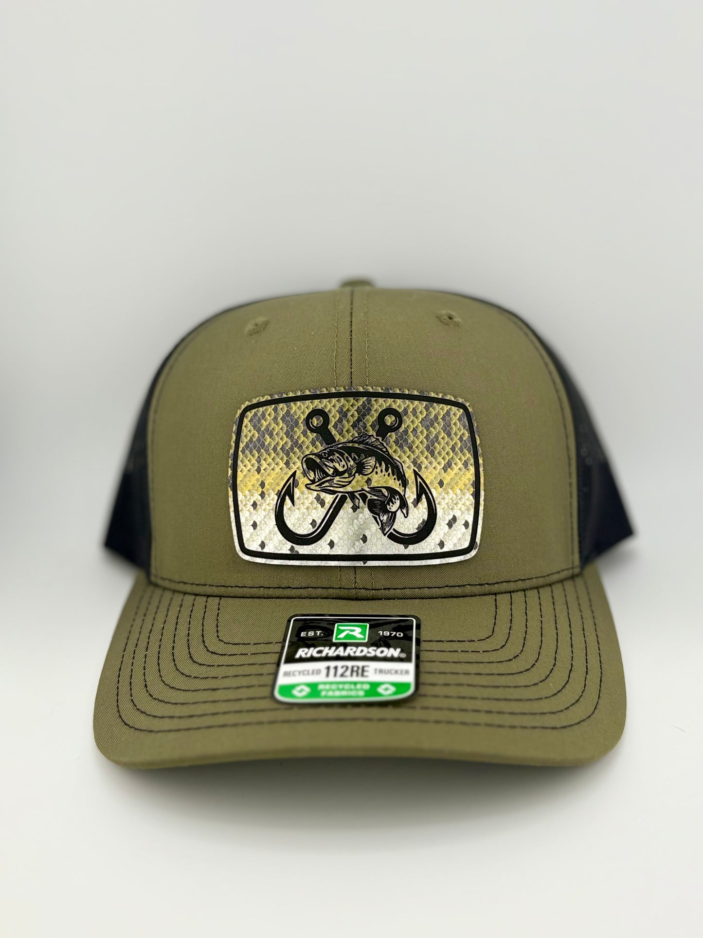 "Olive green and black trucker hat with a structured front panel and a mesh back. The front features a rectangular patch with a fish scale pattern, a detailed illustration of a fish intertwined with double fishing hooks, and black embroidered edges. A Richardson 112RE label is affixed to the brim, indicating its recycled fabric material."