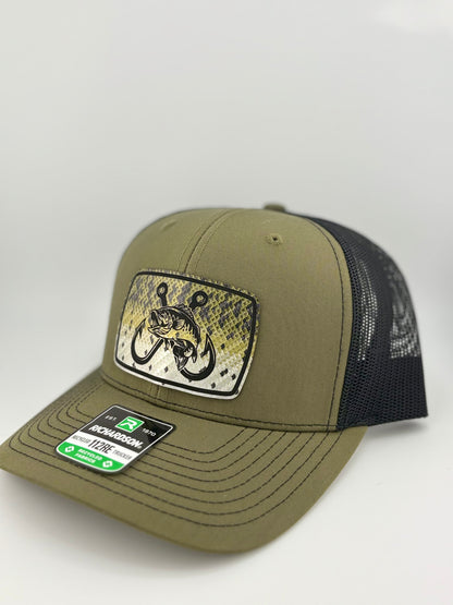 "Side view of an olive green trucker hat with a black mesh back and a structured front panel. The front patch showcases a fish-scale background with a bold fish and double hook design, blending outdoor and fishing aesthetics. The hat's curved brim has visible stitching, and a Richardson 112RE label is placed on it."